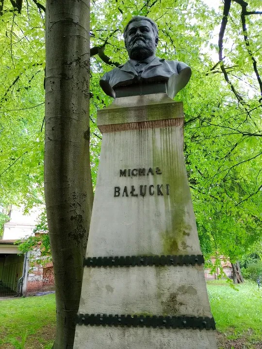 The bust and inscribed base beneath park trees at Michał Bałucki monument