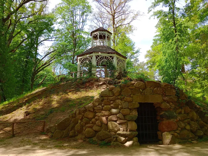 A small open pavilion stands above a stone grotto, reached from the surrounding park paths in Zatonie Ducal Park